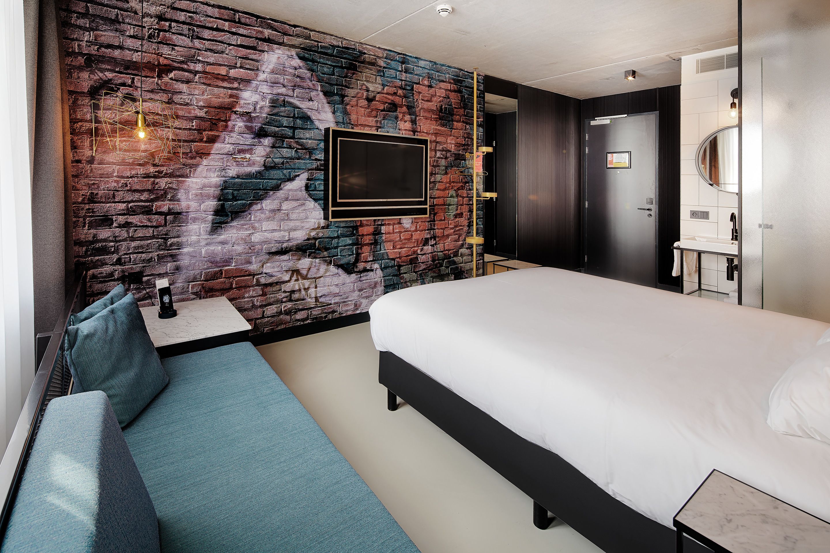 A modern hotel room with a large bed, a seating area, a brick wall with a mural, a mounted TV, and a bathroom with a mirror and sink.