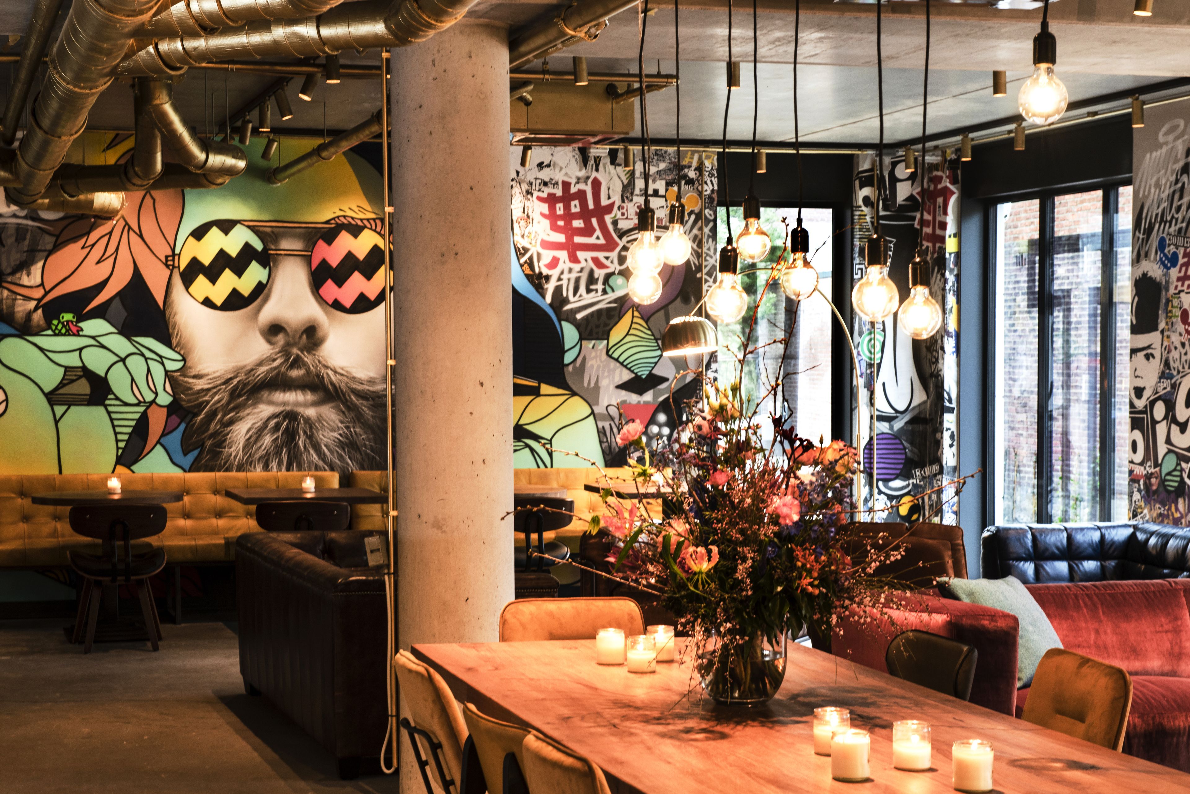 A restaurant with a mural, exposed pipes, hanging light bulbs, and a wooden table with candles.
