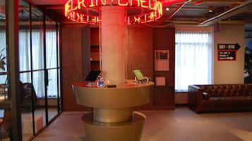 A reception desk with a neon sign, a laptop, and a smartphone on it. There is a couch and a sign indicating parking and toilets.