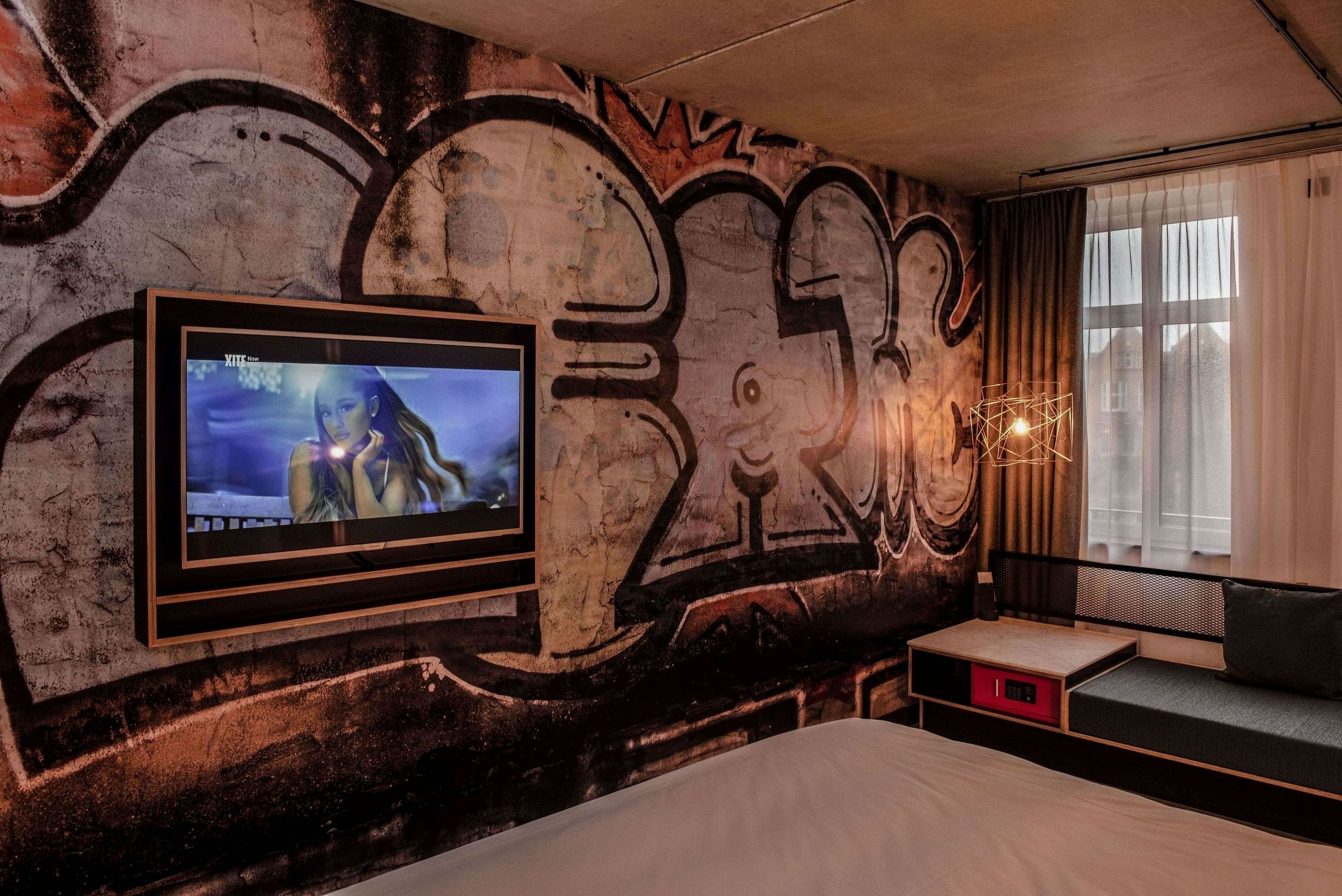 A room with graffiti on the wall, a mounted TV, a bed, and a window with curtains.