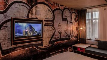 A room with graffiti on the wall, a mounted TV, a bed, and a window with curtains.