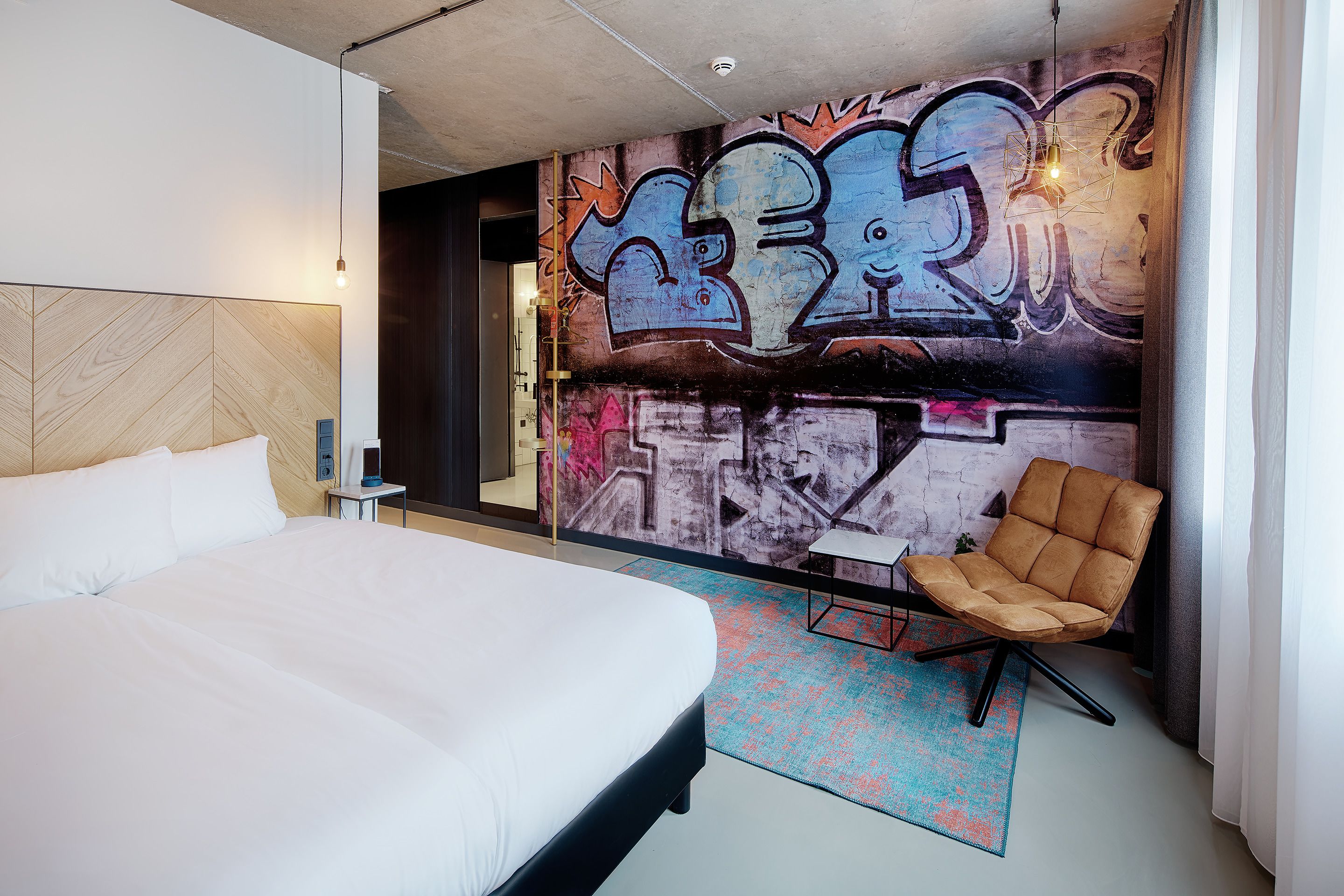 Une chambre d’hôtel moderne avec un grand lit, un coin salon avec un fauteuil et un mur décoré de graffitis.