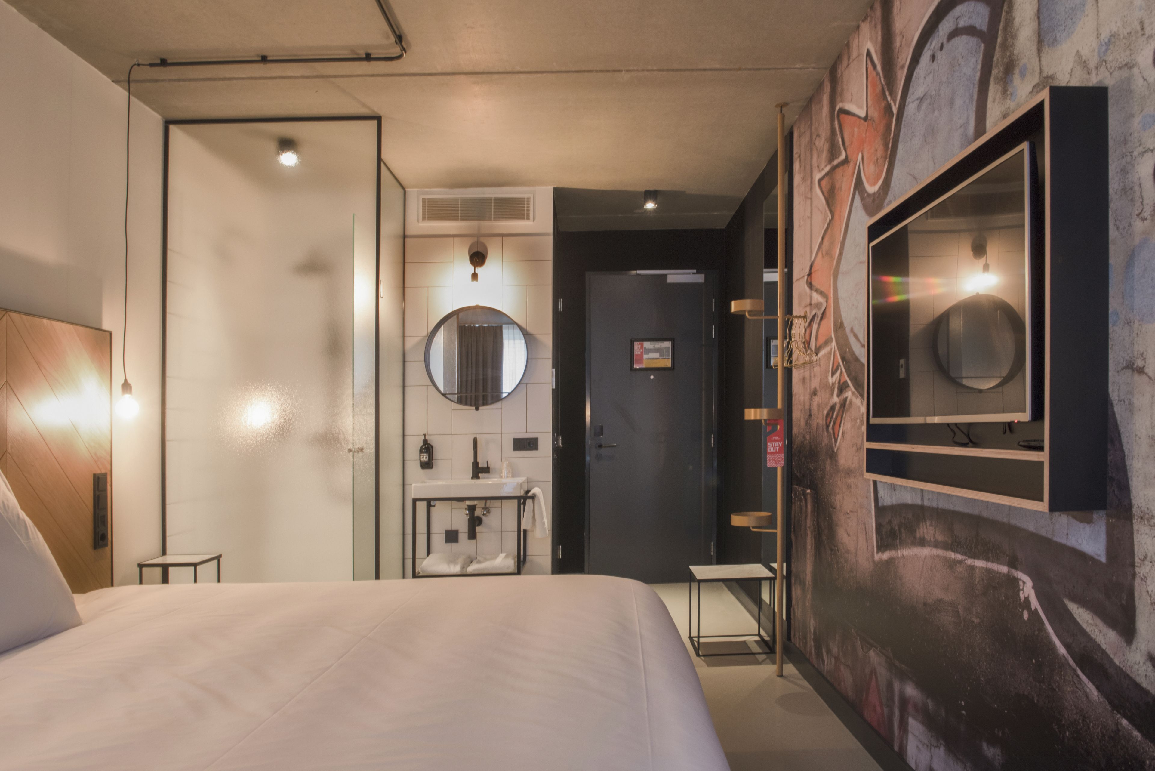 A modern hotel room with a large bed, a round mirror, a small bedside table, a built-in sink, and a wall-mounted TV.