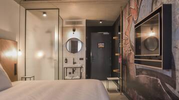 A modern hotel room with a large bed, a round mirror, a small bedside table, a built-in sink, and a wall-mounted TV.
