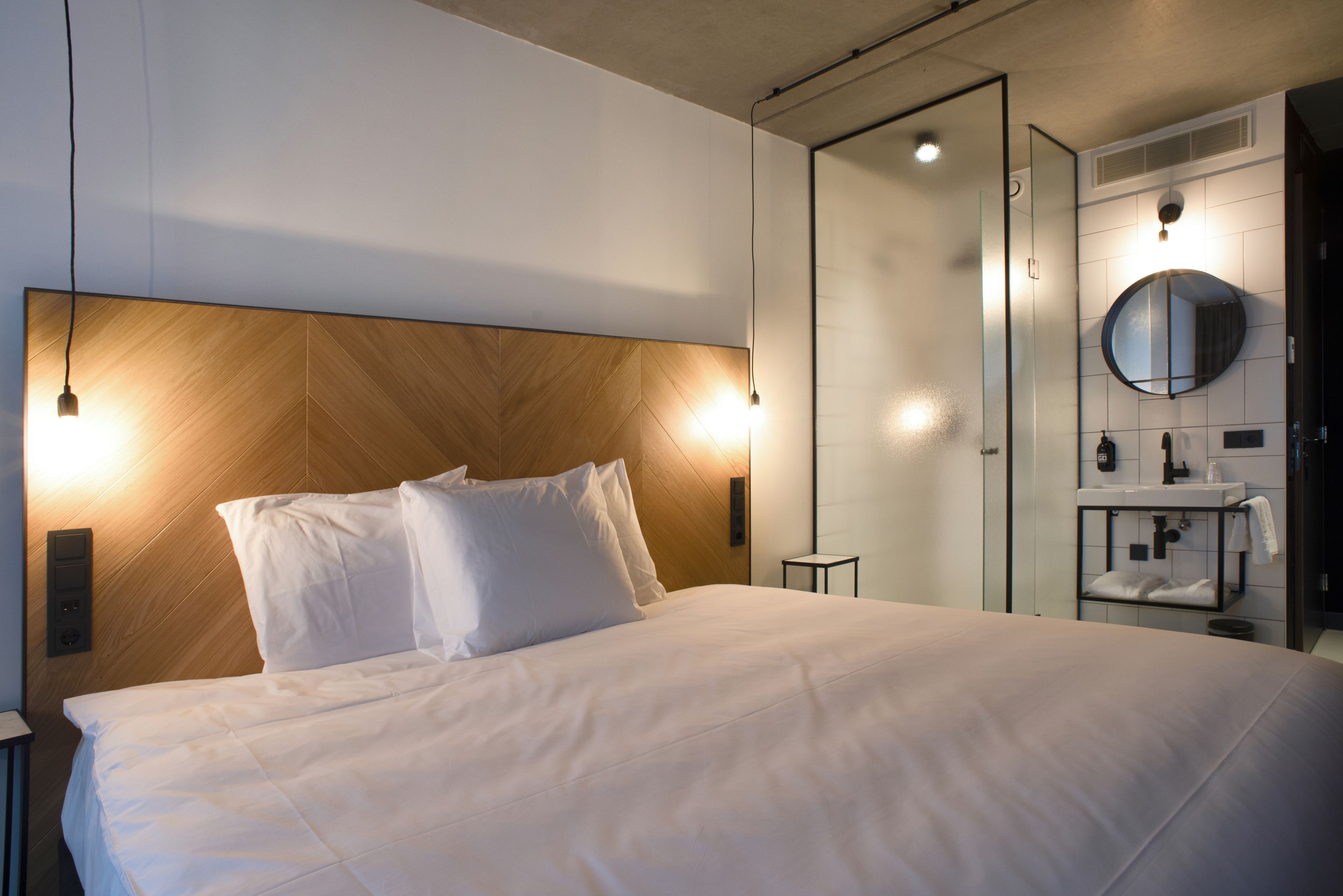 A hotel room with a large bed, a wooden headboard, bedside lamps, a small table, and a bathroom with a mirror and sink.