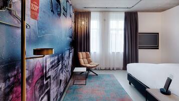 A hotel room with a bed, a chair, a desk, a TV, and a wall with graffiti.