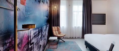 A hotel room with a bed, a chair, a desk, a TV, and a wall with graffiti.