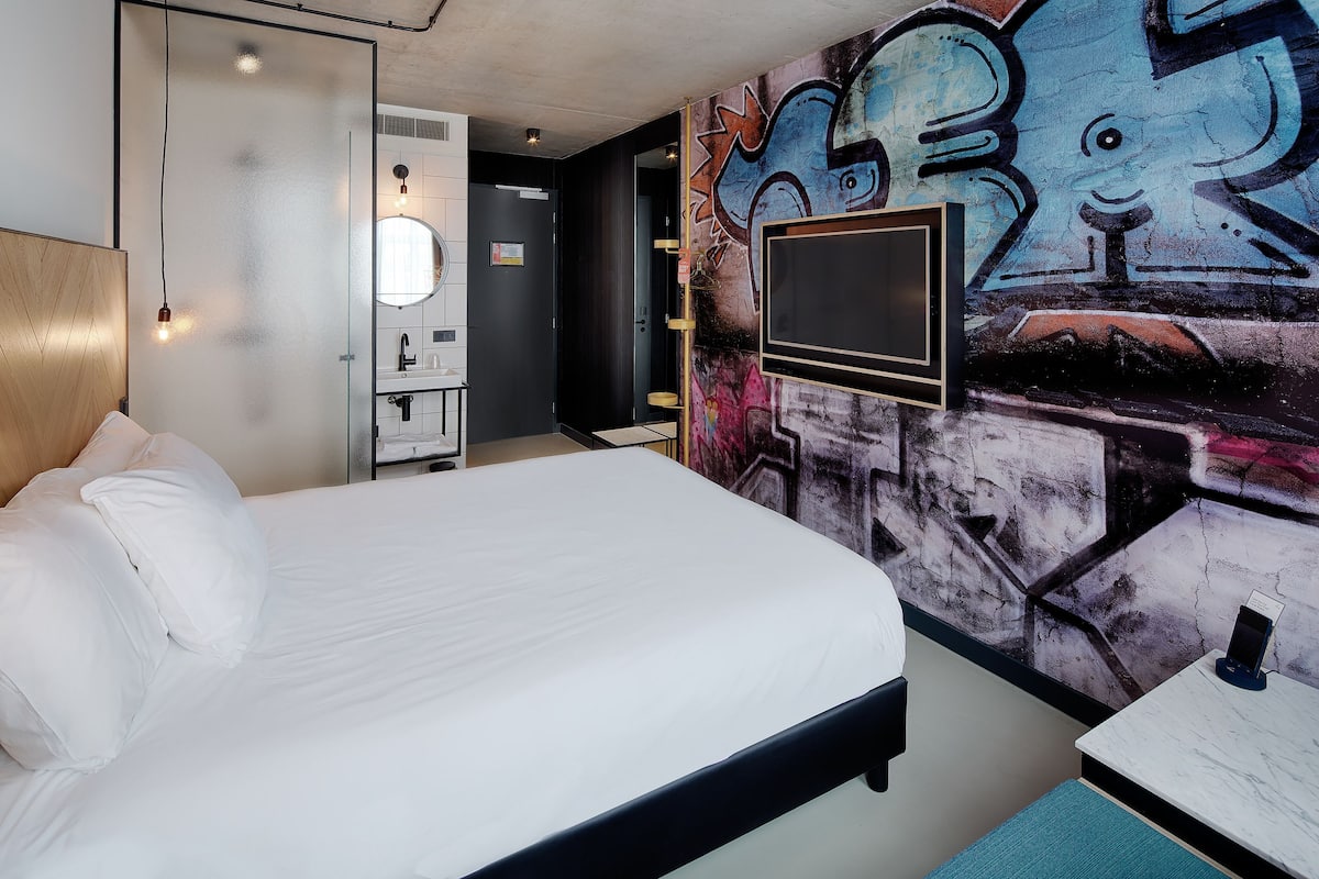 Una habitación de hotel moderna con una cama grande, un televisor montado en la pared y una obra de arte callejero.