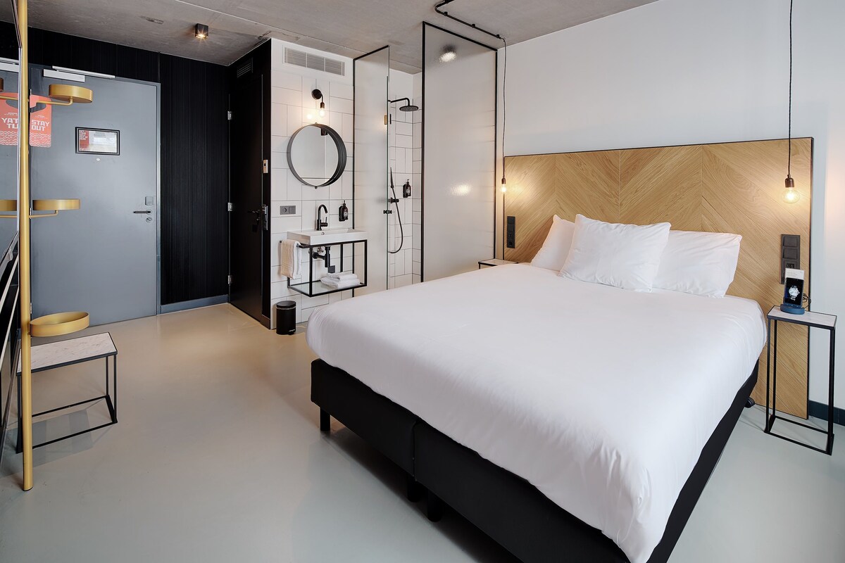 A modern hotel room with a large bed, a shower, a vanity with a mirror, and a small table.