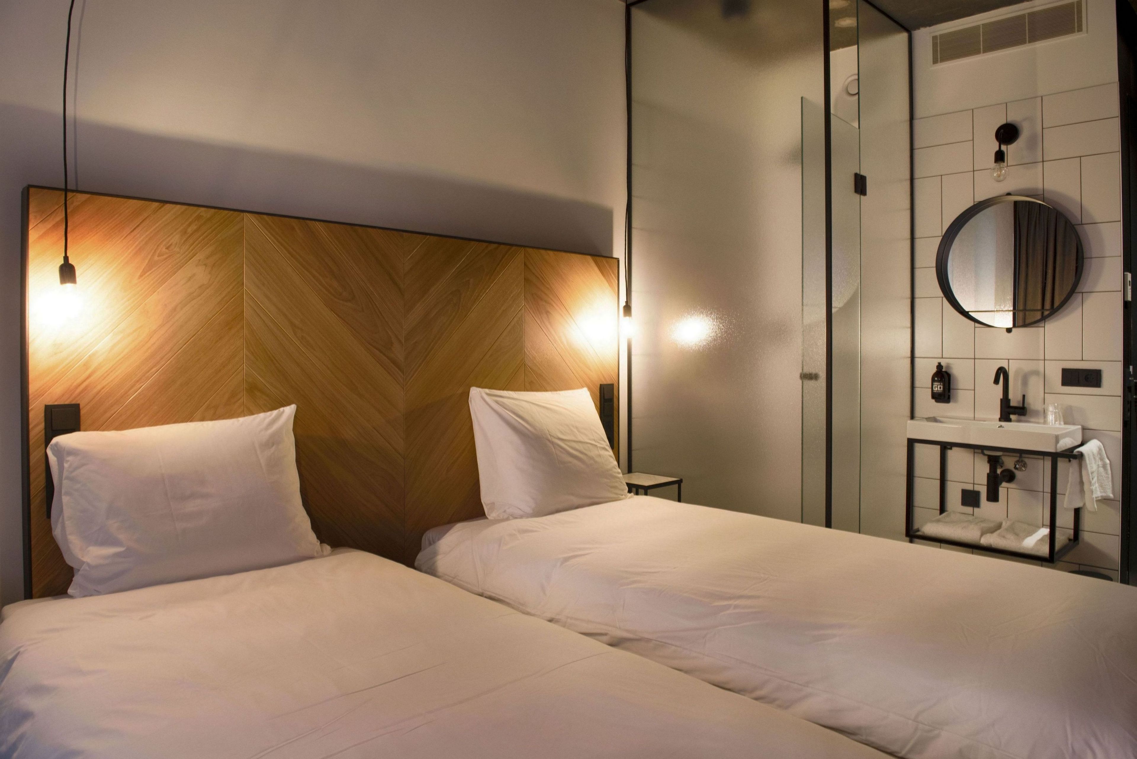 Ein Hotelzimmer mit einem hölzernen Kopfteil, zwei Betten, einem runden Spiegel, einem Waschbecken und einem Handtuchhalter.