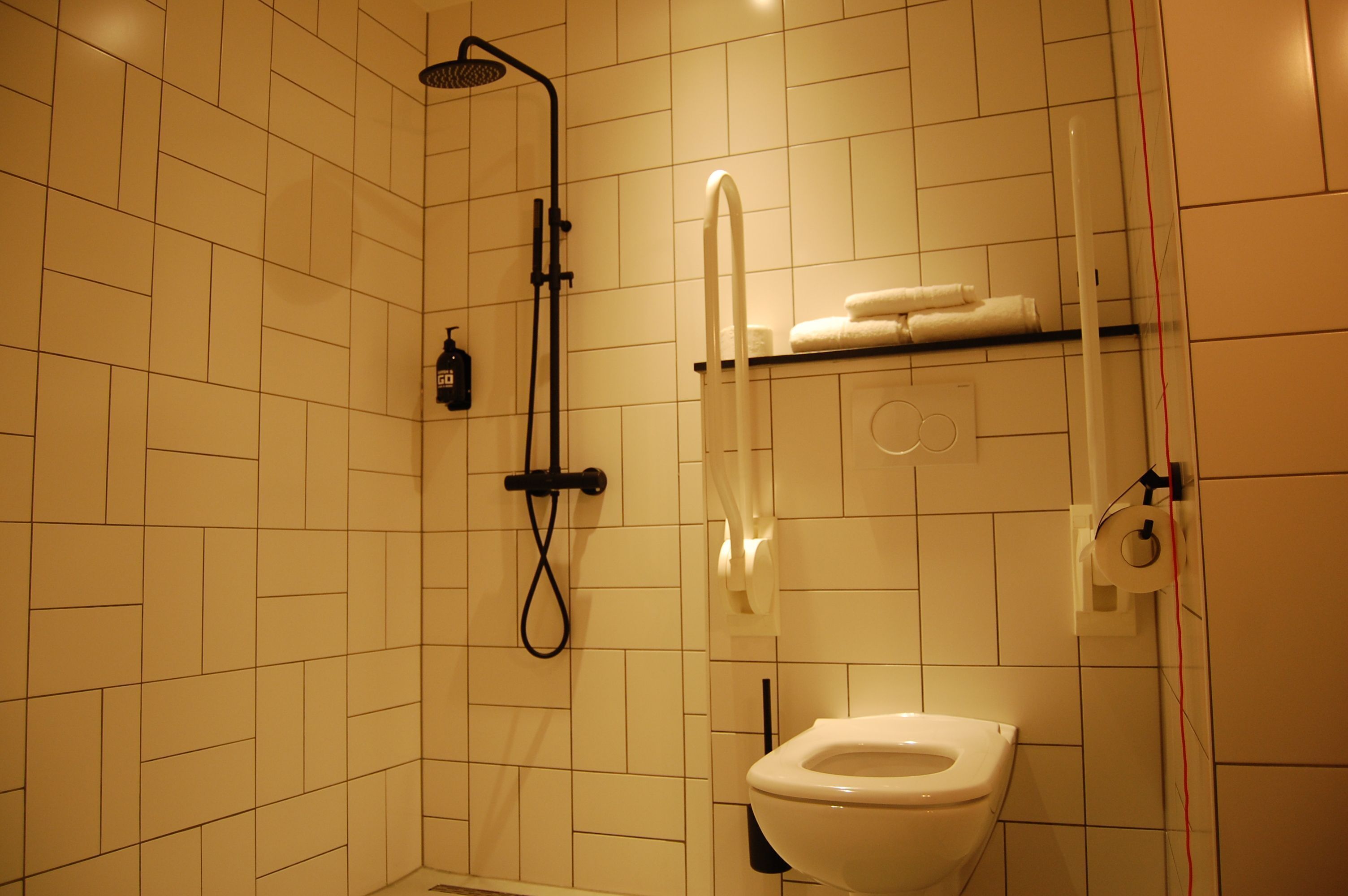 Une salle de bain avec des carreaux blancs, une douche, des toilettes et un porte-serviettes.