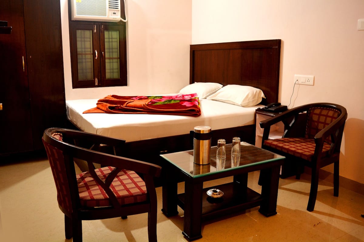 A hotel room with a bed, a nightstand, two chairs, a small table with a thermos, glasses, and a remote control.