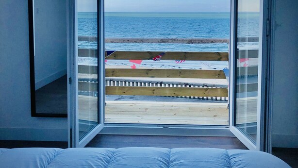 Un dormitorio con una cama grande, vista a la playa a través de una puerta abierta y una terraza de madera con banderas de playa.