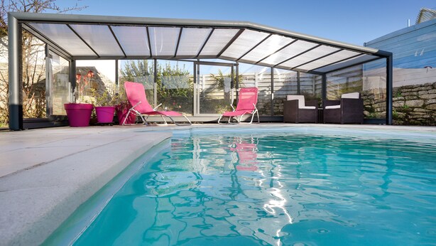 A swimming pool with a glass enclosure, pink chairs, and potted plants.