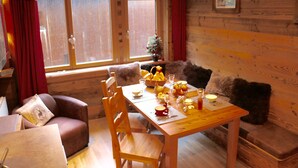 Interior acogedor de una cabaña con una mesa de comedor de madera dispuesta para cuatro personas, rodeada de sillas de madera. Se ve un pequeño árbol de Navidad y un cuenco de fruta sobre la mesa.
