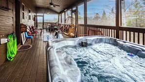 Un bain à remous sur une terrasse en bois, avec des chaises et une table, de grandes fenêtres et une vue sur les arbres.