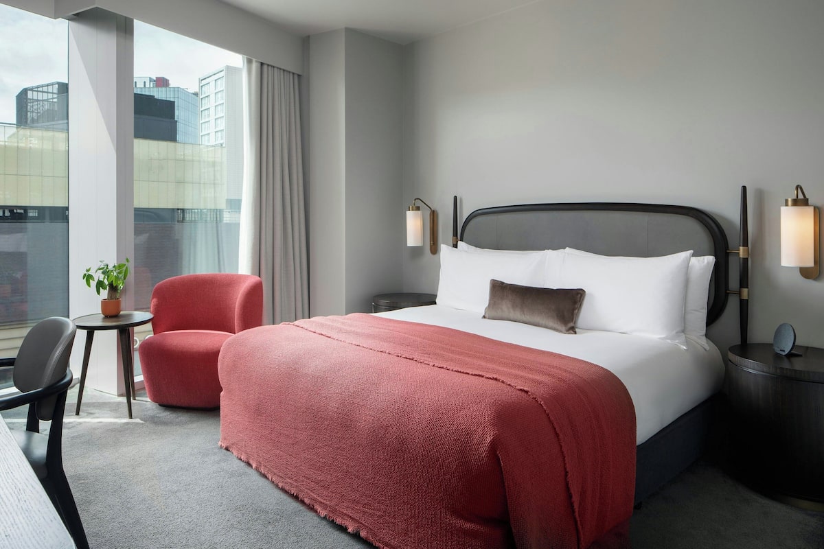 Hotellihuone, jossa on suuri sänky, kaksi yöpöytävalaisinta, punainen tuoli, pieni pöytä, jossa on kasvi, ja suuret ikkunat, joista on näkymä rakennuksiin.