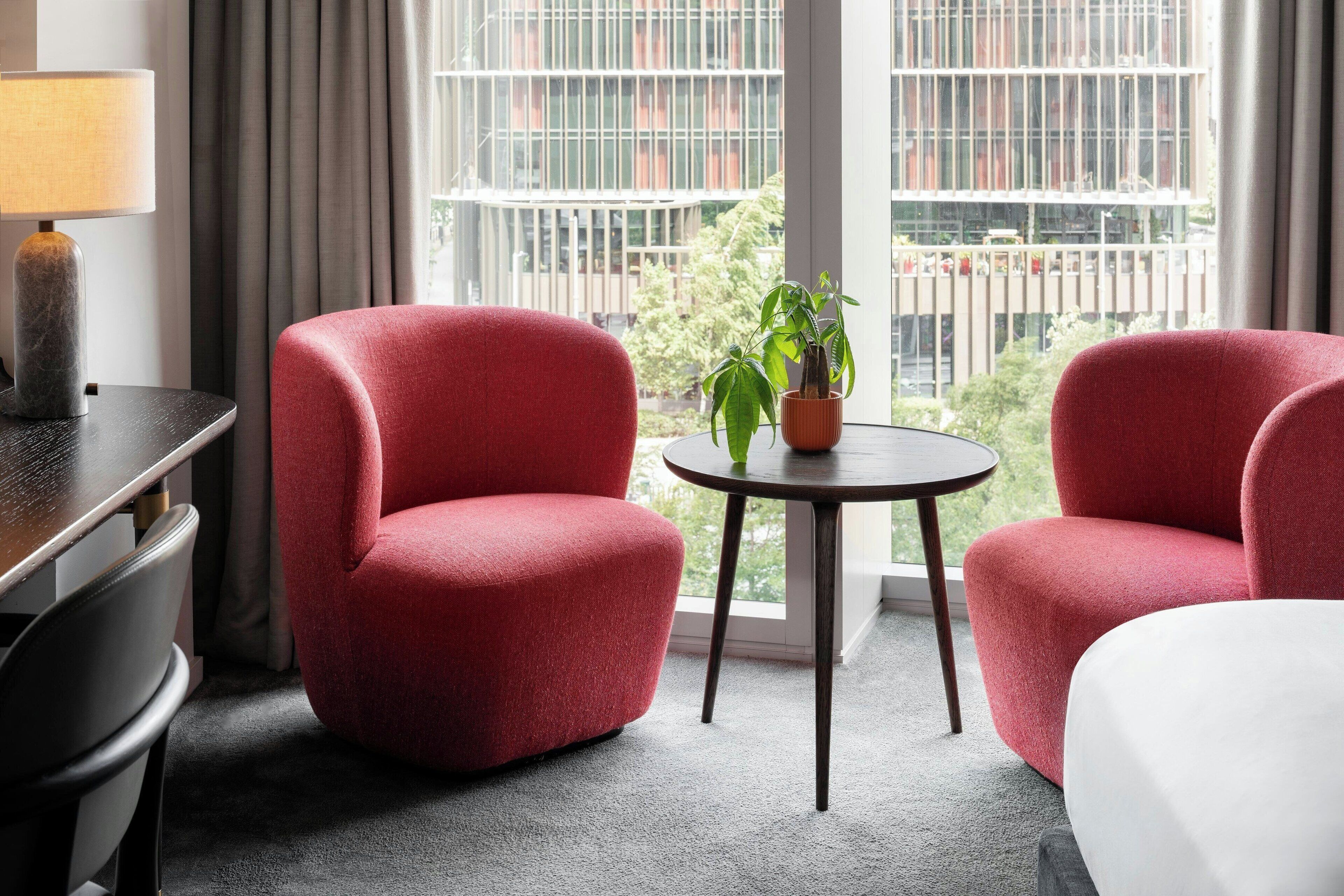 Ein Hotelzimmer mit zwei roten Sesseln, einem kleinen runden Tisch mit einer Topfpflanze, einer Lampe und Blick auf ein Gebäude durch große Fenster.