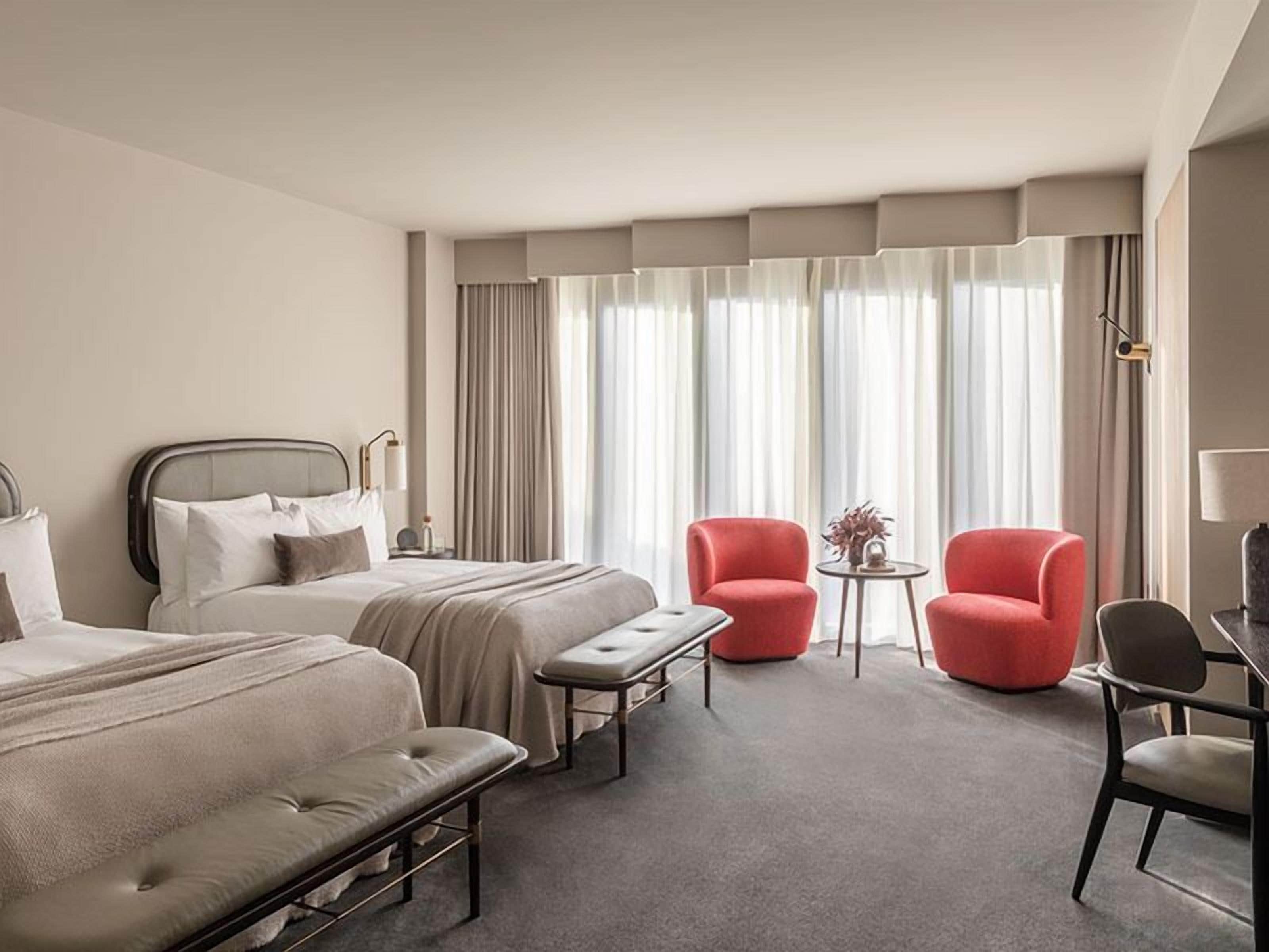 A hotel room with two beds, a bench, two red chairs, a small table, and a lamp.