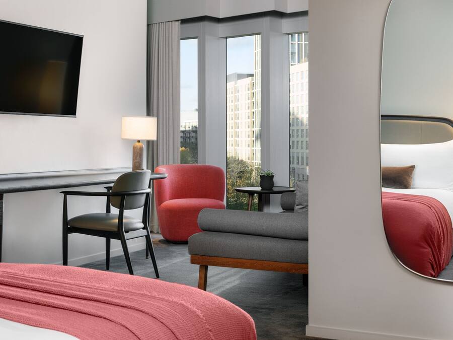 A hotel room with a bed, a desk, a chair, a red armchair, a grey sofa, a small table with a lamp, and a large mirror.