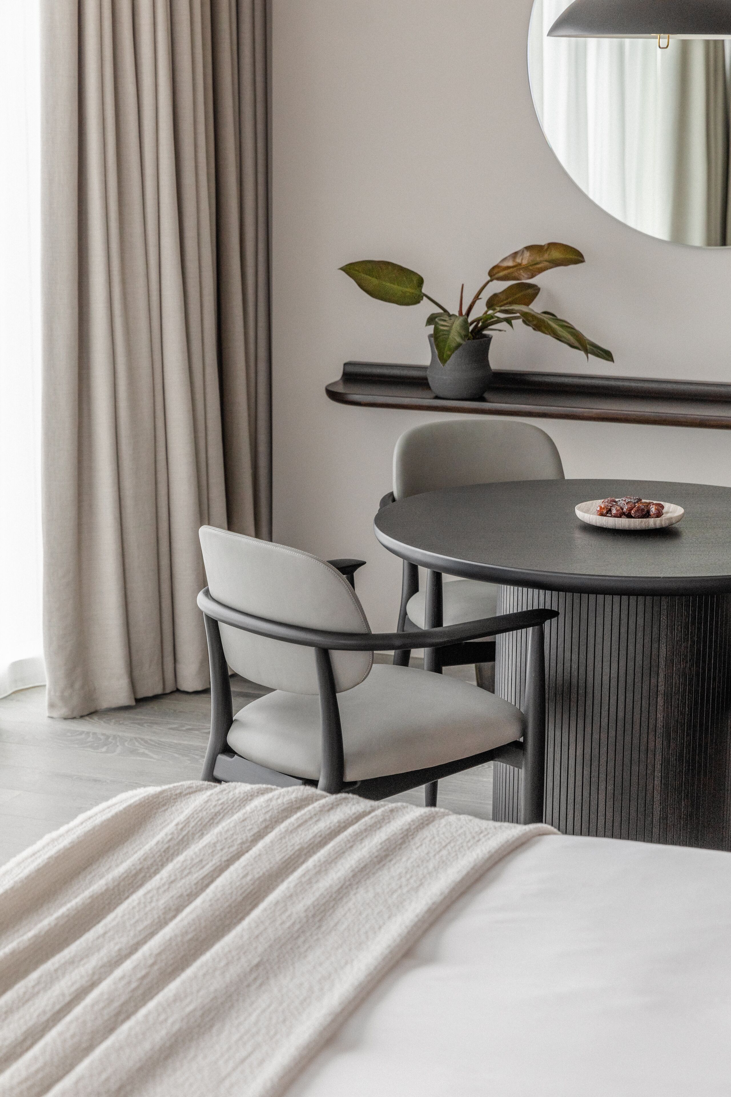 Una habitación moderna con una mesa de comedor redonda, dos sillas, una planta en maceta, un espejo y una cama con una manta blanca.