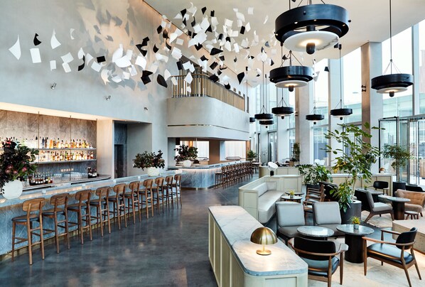 A modern bar area with a long counter, multiple seating options, and a high ceiling with hanging light fixtures. - The Stratford, Autograph Collection (London)