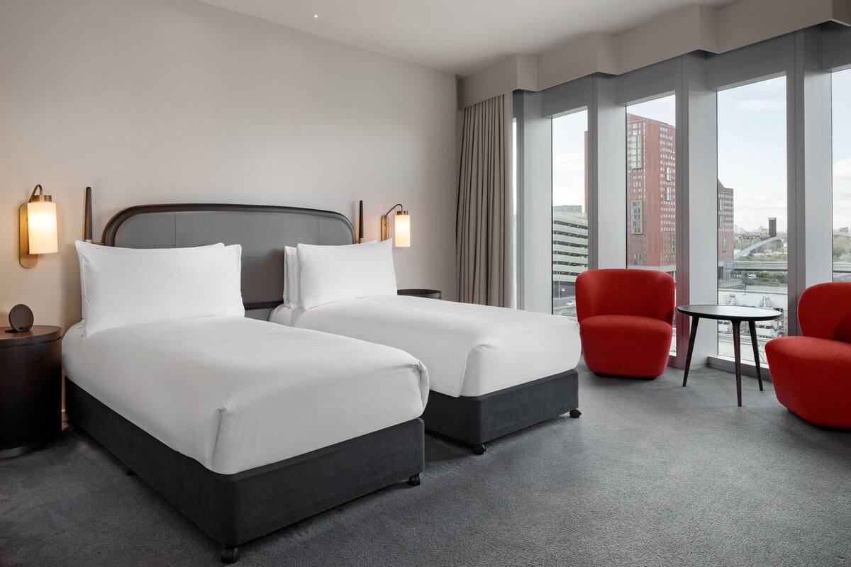 Hotellihuone, jossa on kaksi sänkyä, punainen nojatuoli, pieni pöytä ja suuri ikkuna, josta avautuu näkymä kaupunkiin.