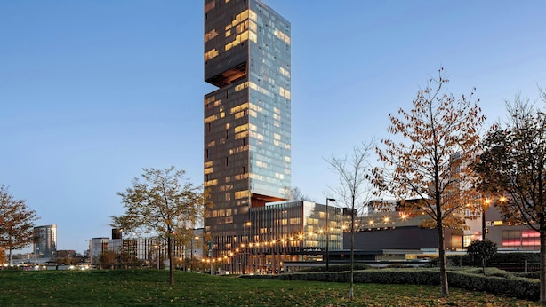 Un edificio moderno de gran altura con un diseño arquitectónico singular, rodeado por un parque bien cuidado con árboles y senderos.