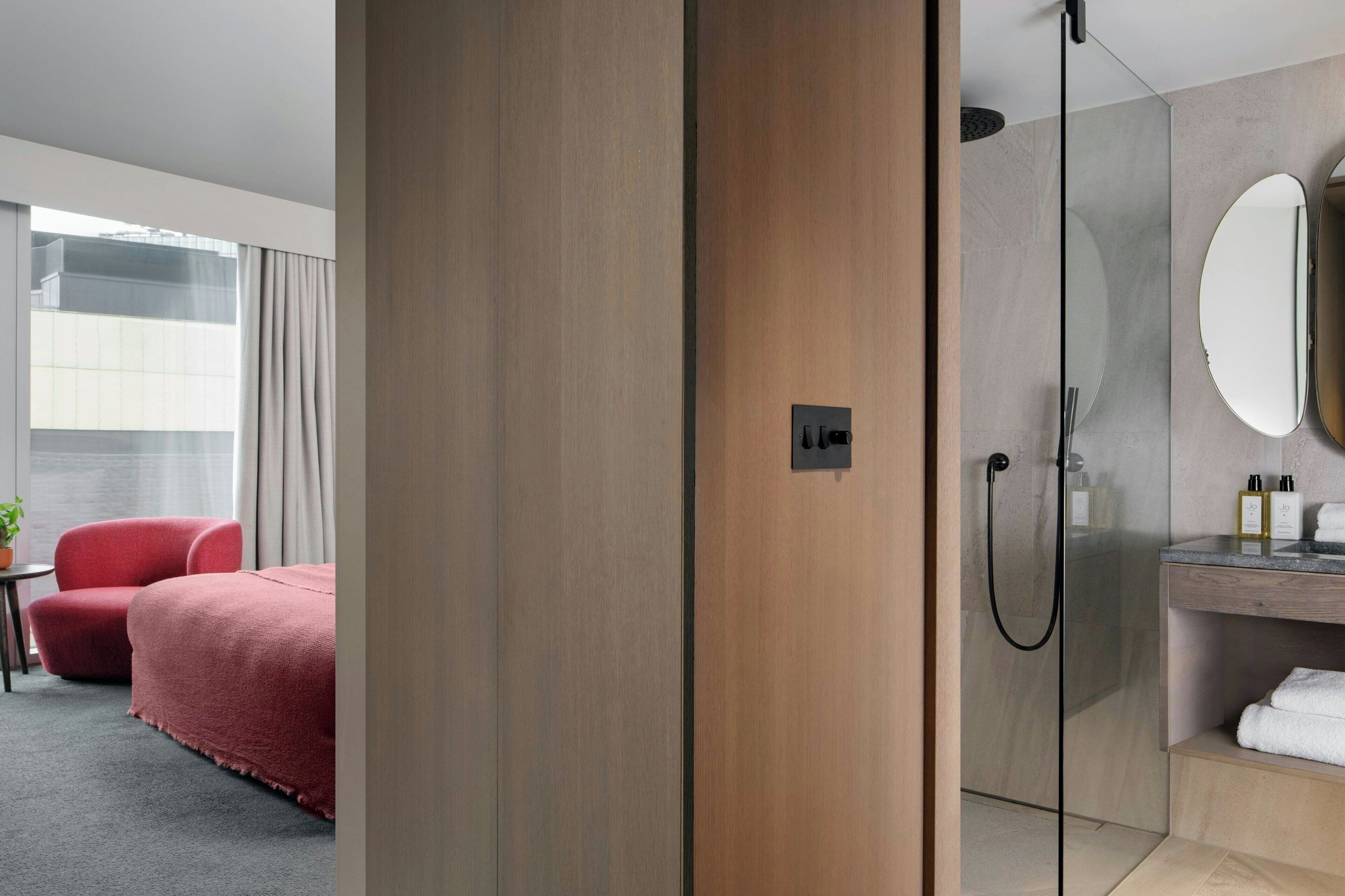 Habitación de hotel con cama, sillón rojo, cabina de ducha de vidrio y baño con espejo y artículos de aseo.