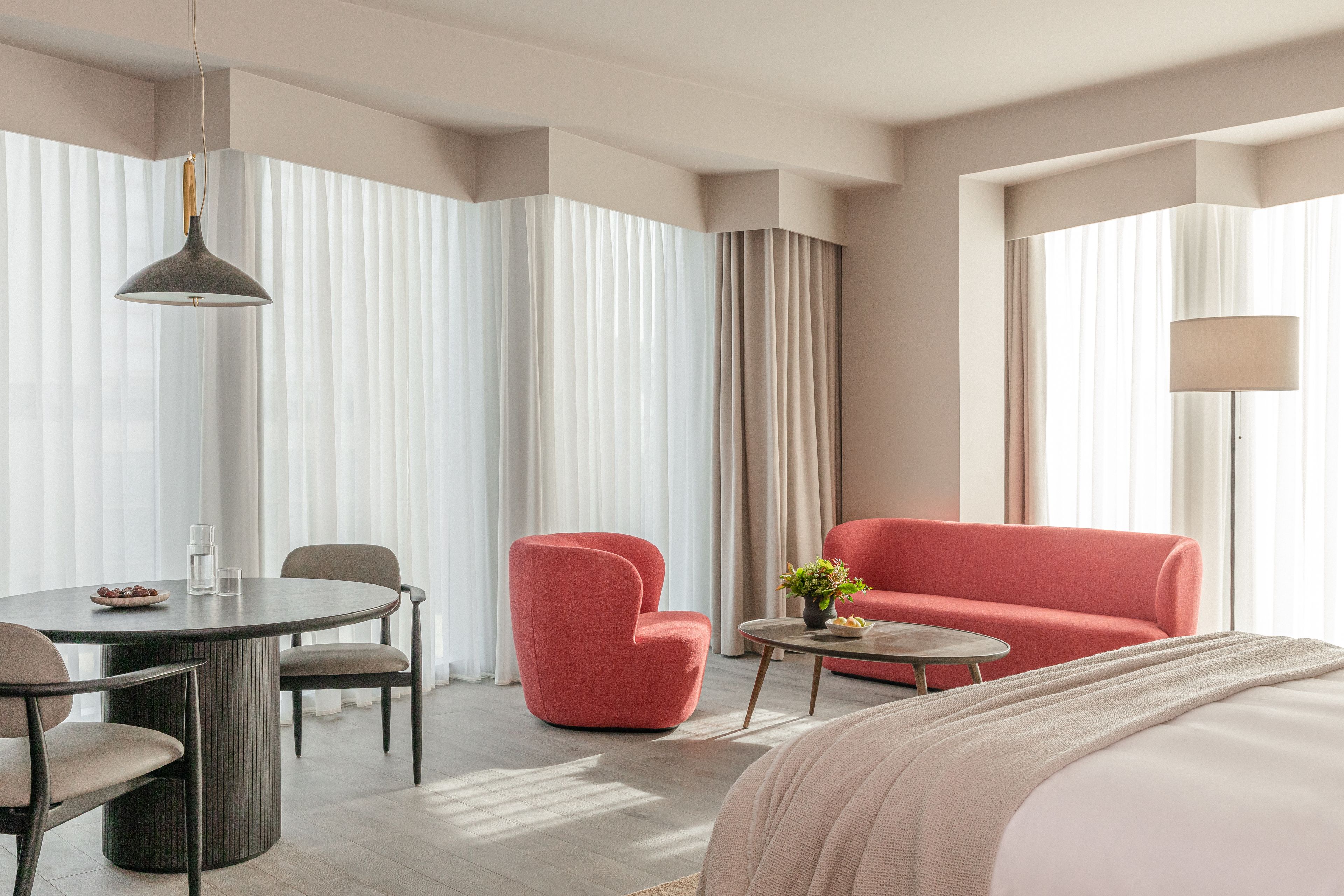 Ein Hotelzimmer mit einem runden Esstisch, zwei Stühlen, einem roten Sessel, einem kleinen Beistelltisch mit Pflanze, einem Sofa und einem Bett mit weißem Kopfteil.