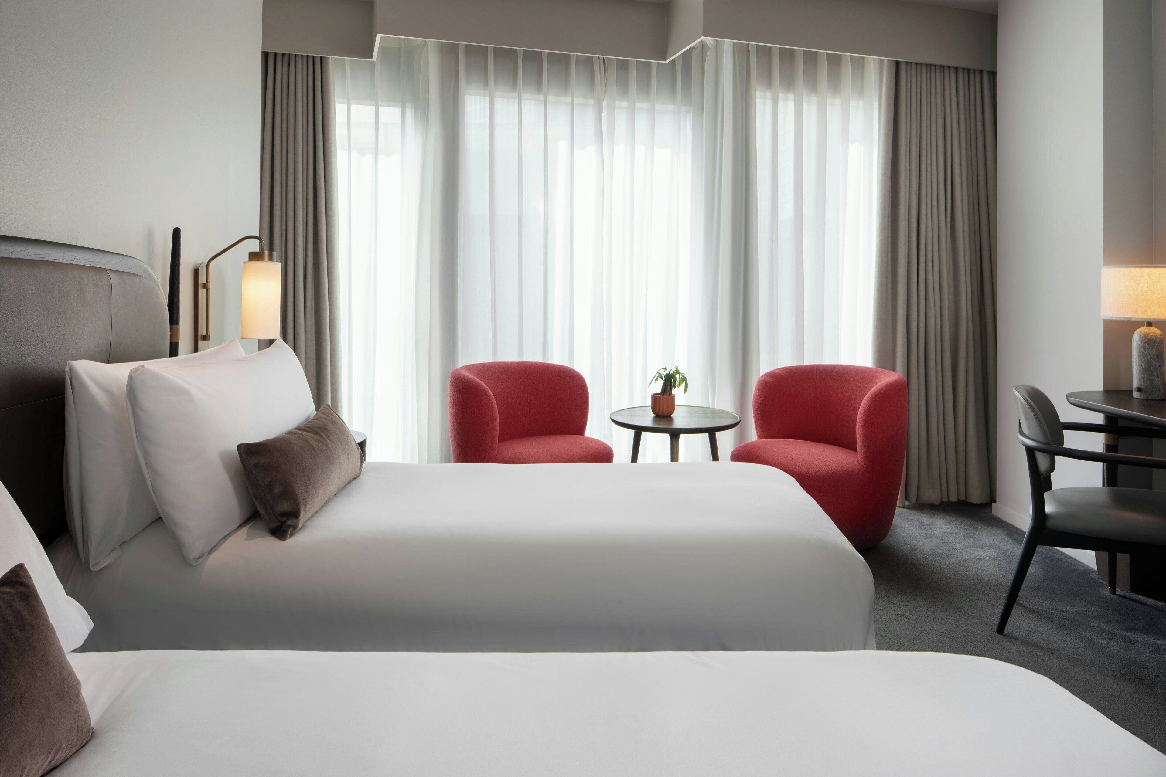 Une chambre d’hôtel avec un grand lit, deux fauteuils rouges, une petite table ronde avec une plante et un bureau avec une lampe.