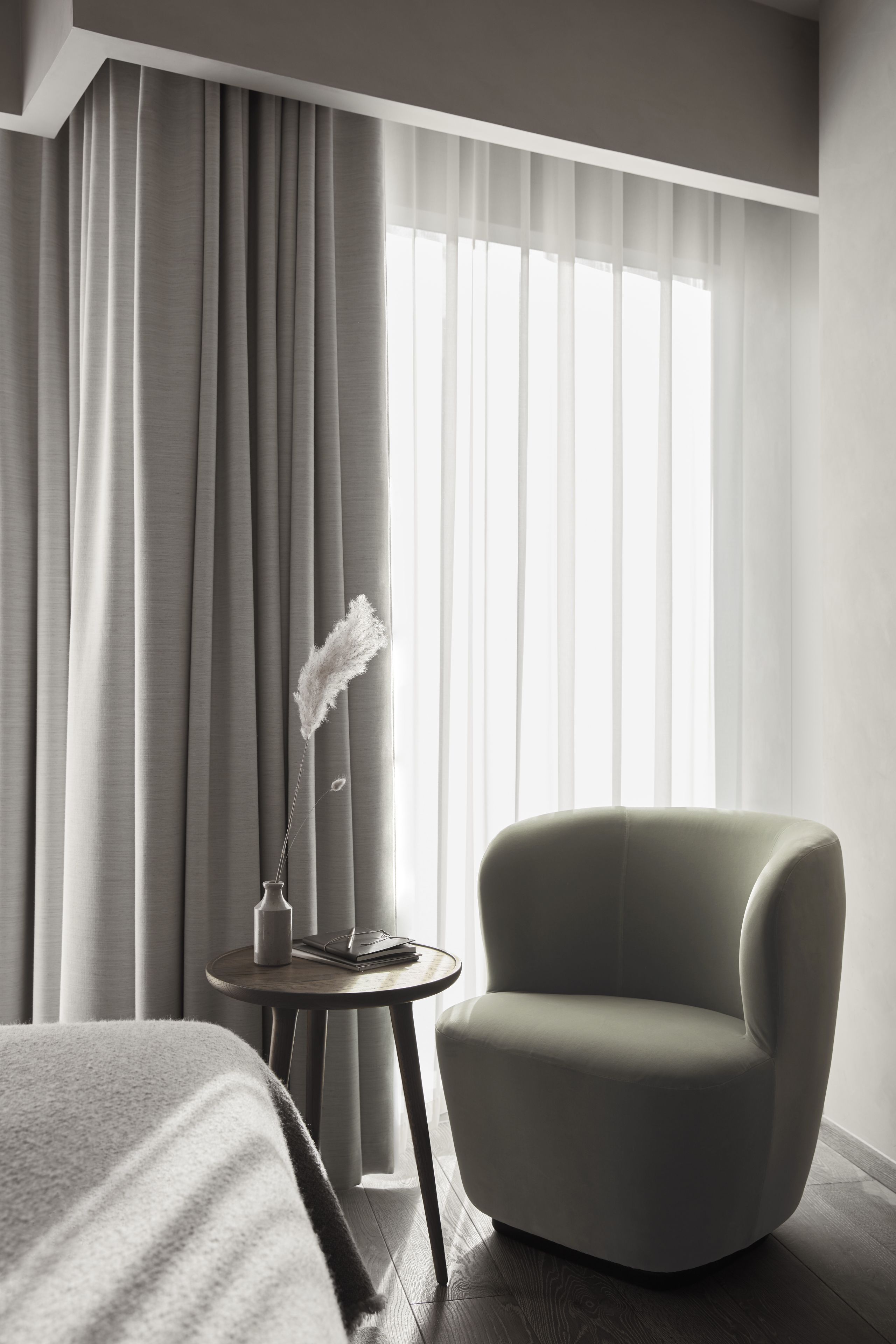 Une chambre d’hôtel moderne avec une grande fenêtre, un fauteuil gris, une petite table ronde ornée d’un vase et d’un livre, et un lit recouvert d’une couverture à rayures.