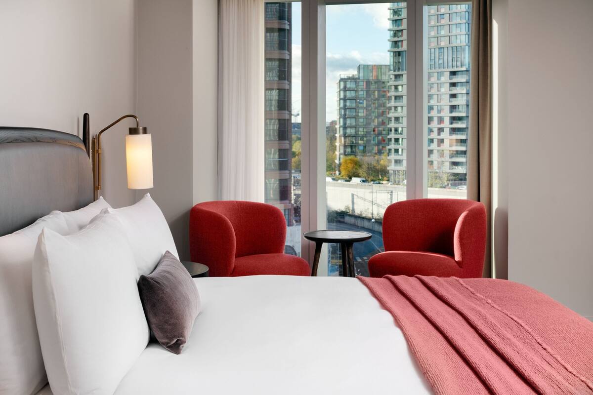 Hotellihuone, jossa on sänky, kaksi punaista nojatuolia, pieni pyöreä pöytä ja suuret ikkunat, joista avautuu näkymä korkeisiin rakennuksiin.