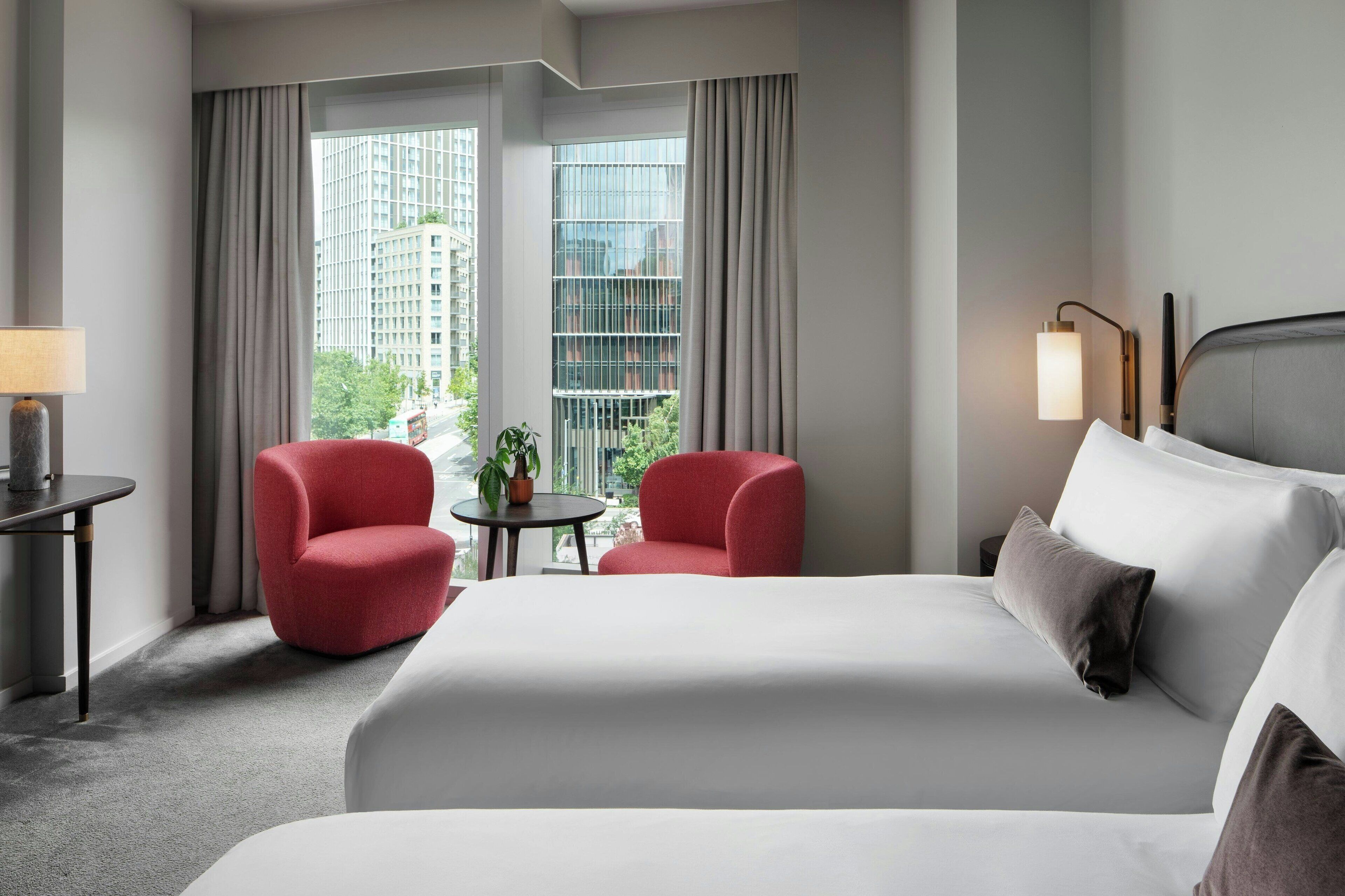 Habitación de hotel con una cama grande, dos sillones rojos, una mesita con una planta y vistas a edificios a través de las ventanas.