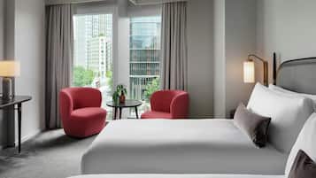 Habitación de hotel con una cama grande, dos sillones rojos, una mesita con una planta y vistas a edificios a través de las ventanas.