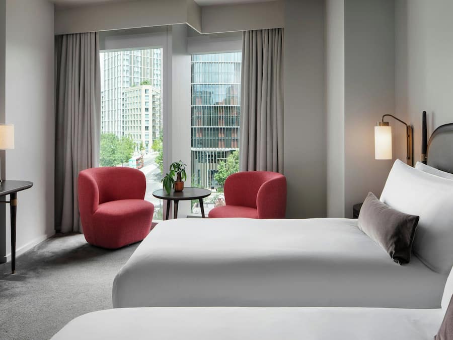 Habitación de hotel con una cama grande, dos sillones rojos, una mesita con una planta y vistas a edificios a través de las ventanas.