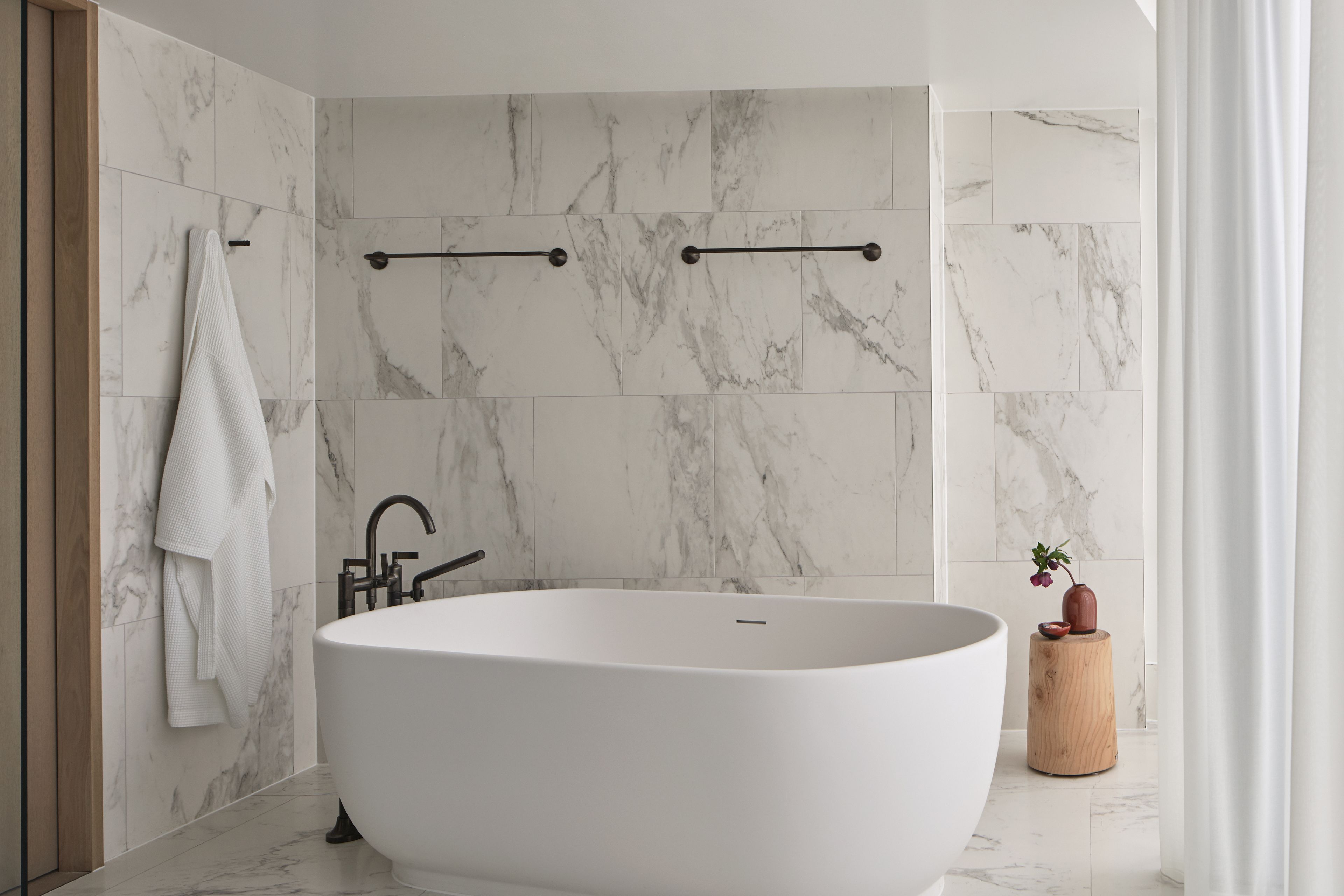 Ein modernes Badezimmer mit einer freistehenden Badewanne, Marmorwänden und einem hölzernen Hocker mit einer Vase.