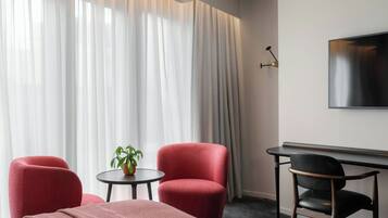 Quarto de hotel com uma cama, duas poltronas vermelhas, uma mesa redonda pequena com uma planta, uma escrivaninha e uma TV fixada na parede.