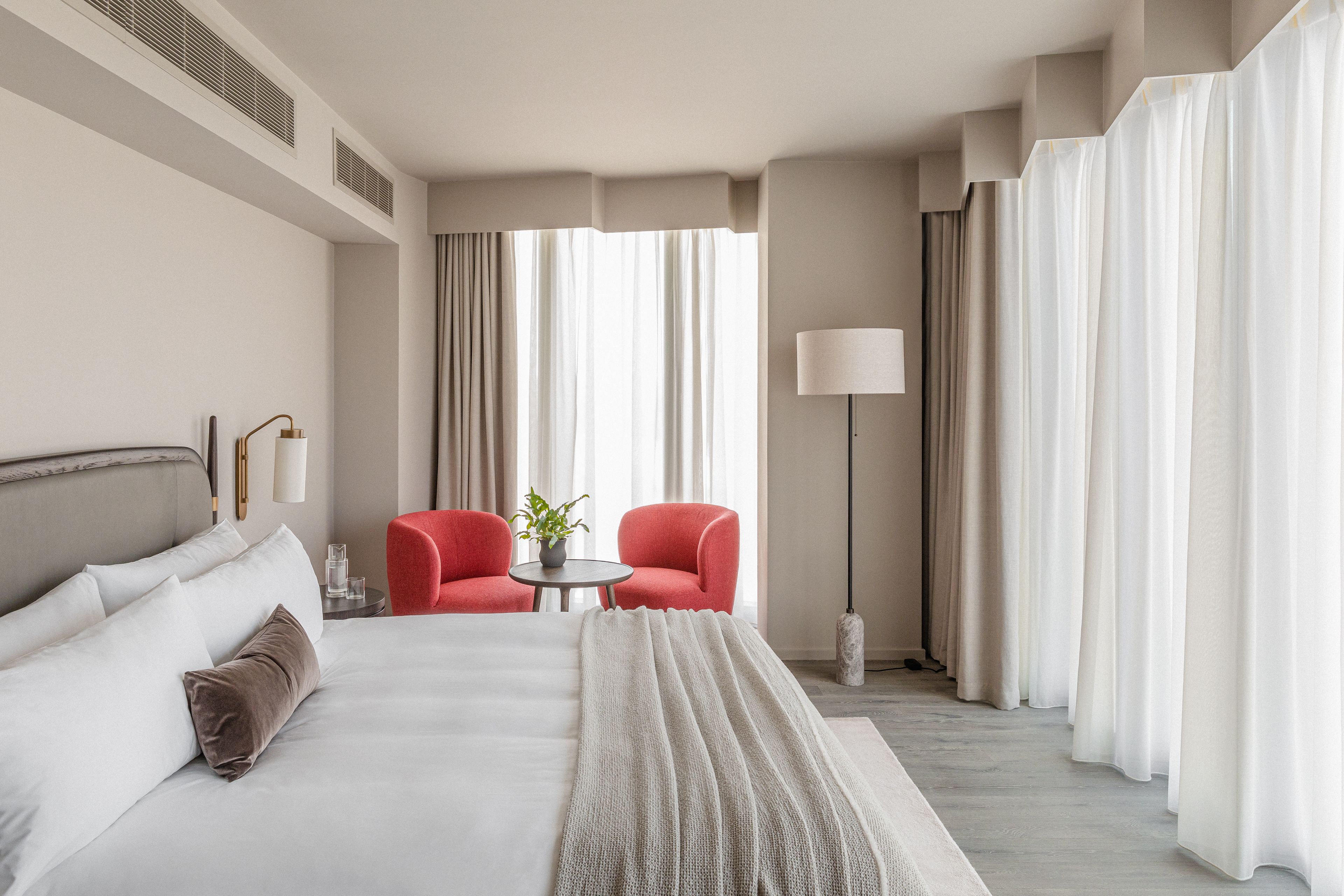 Une chambre d’hôtel avec un grand lit, deux fauteuils rouges, une petite table avec une plante et un lampadaire.