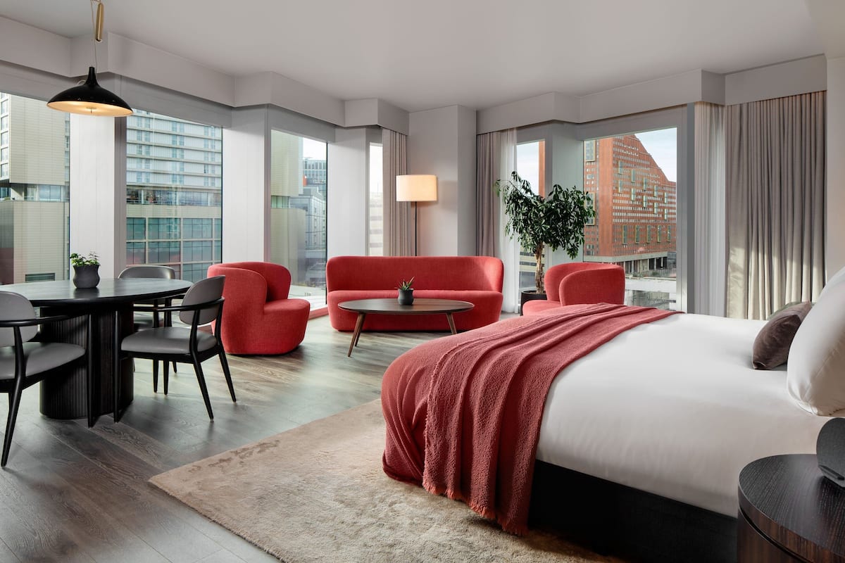 Moderni hotellihuone, jossa on suuri sänky, punaiset nojatuolit, pyöreä sohvapöytä sekä ruokailutila, jossa on pöytä ja tuolit.
