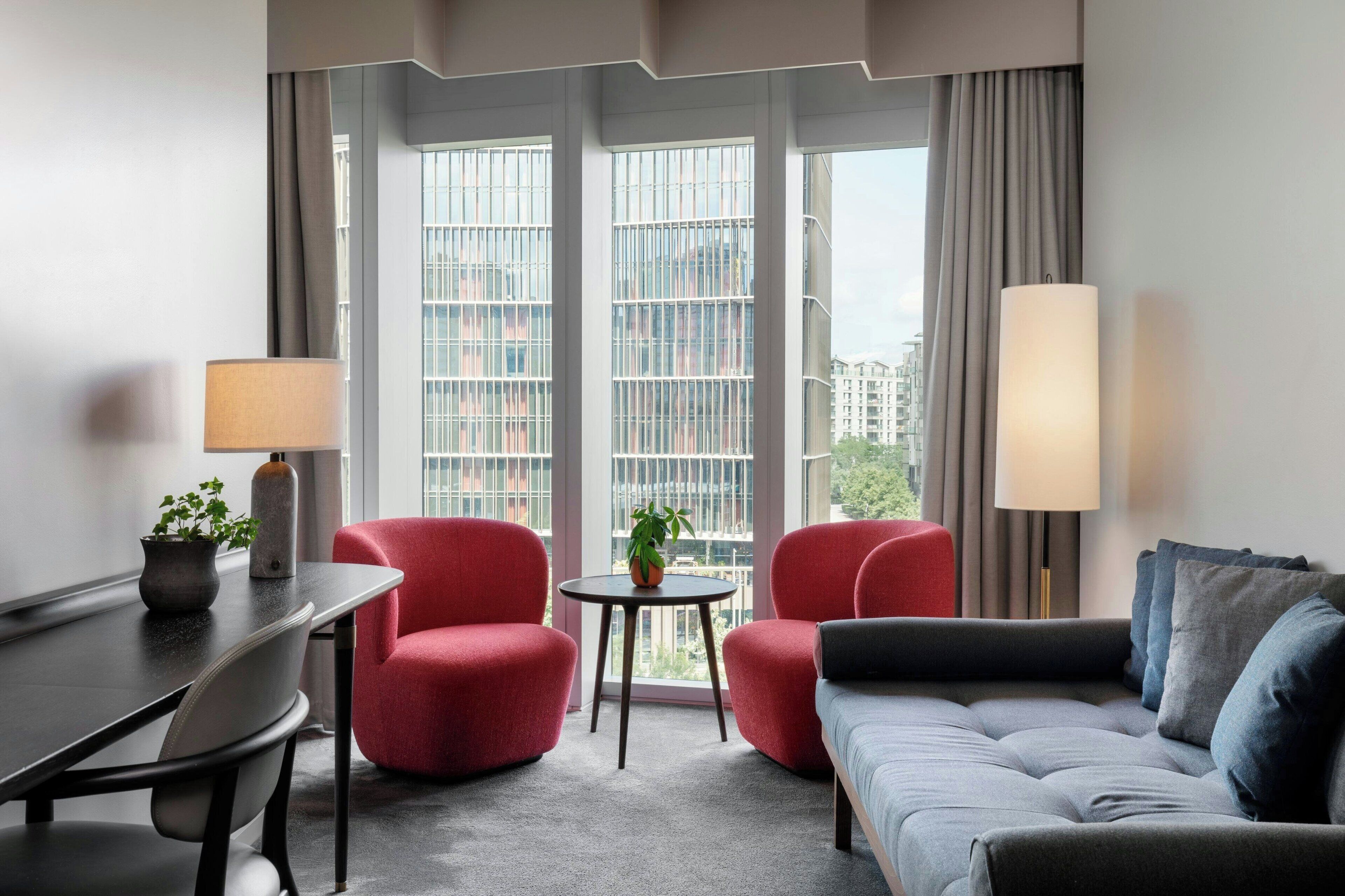 A modern hotel room with a grey sofa, red armchairs, a small round table with a plant, and a lamp. Large windows offer a view of the cityscape.