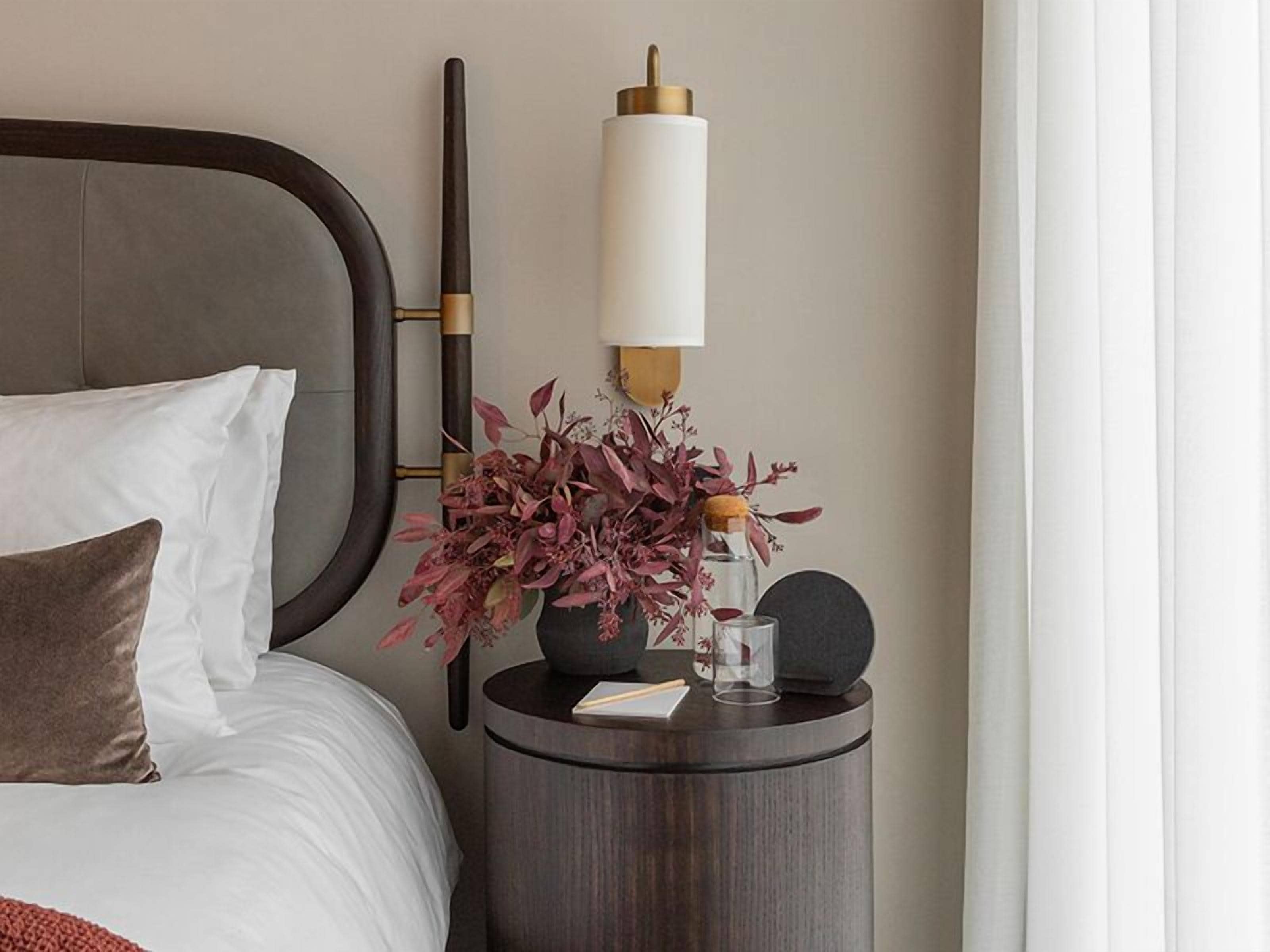 Ein ordentlich bezogenes Bett mit weißen und braunen Kissen, ein runder Nachttisch mit Lampe, eine Vase mit Trockenblumen und ein kleines Buch.