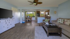 A spacious living area with a television, dining table, and sofa. A ceiling fan is visible, and the room has a view of the outdoors. - Beautiful views of the mountains and oceans on the ground floor (Princeville)