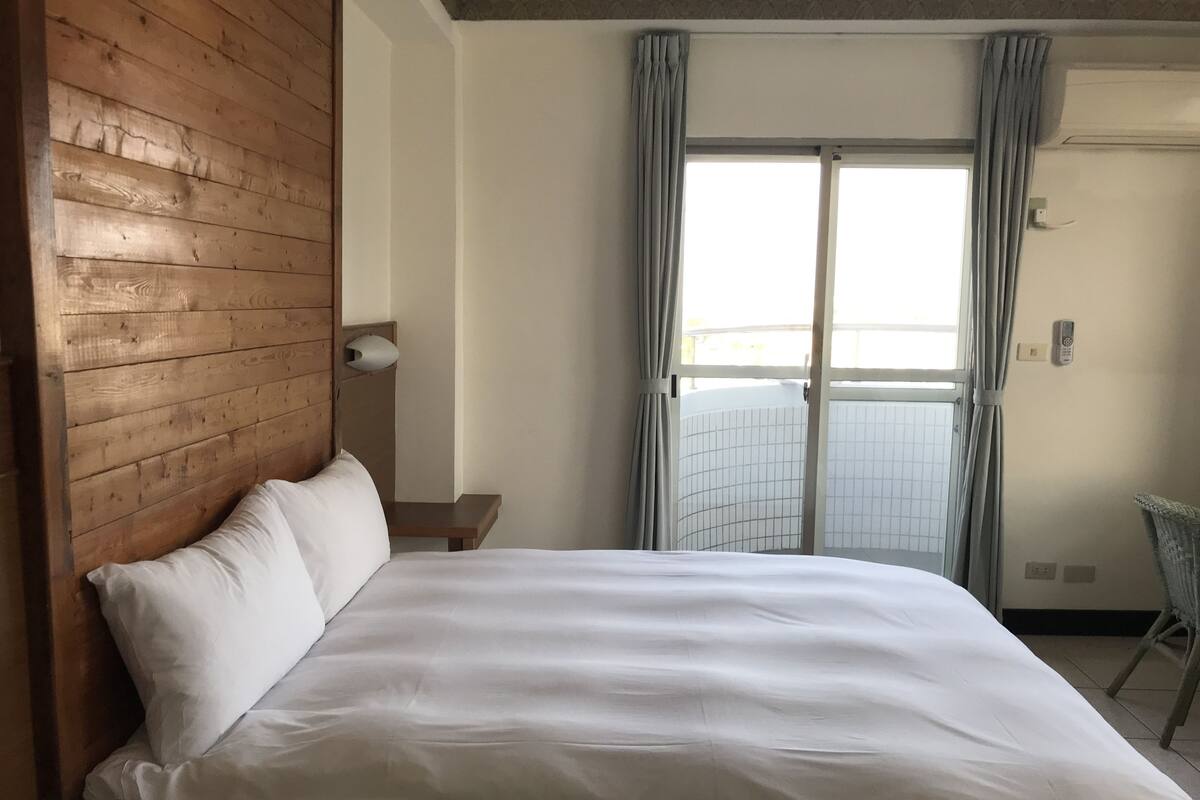 A hotel room with a wooden headboard, a large bed with white linens, a small bedside table, a wall-mounted lamp, a window with curtains, and a view of a balcony.