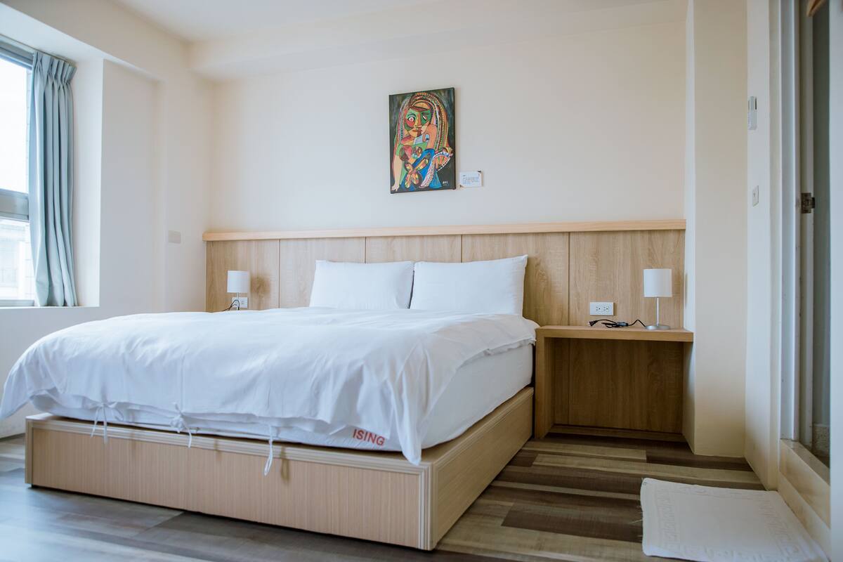 A modern hotel room with a wooden bed, white bedding, a nightstand with a lamp, and a painting on the wall.