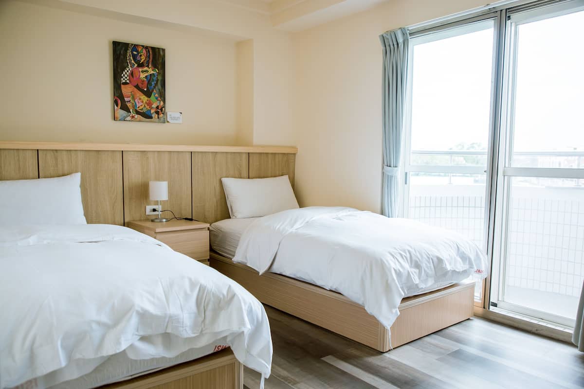 A hotel room with two beds, a nightstand, a painting on the wall, and a large window with curtains.
