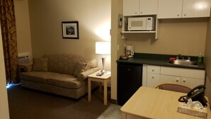 A compact living space with a sofa, a small table with a lamp, a kitchenette with a microwave, and a refrigerator. - Shemron Suites Hotel (Deep River)