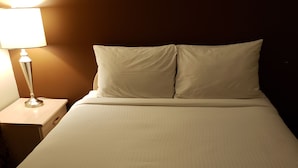 A neatly made bed with white linens and pillows, a bedside lamp, and a wooden nightstand. - Shemron Suites Hotel (Deep River)