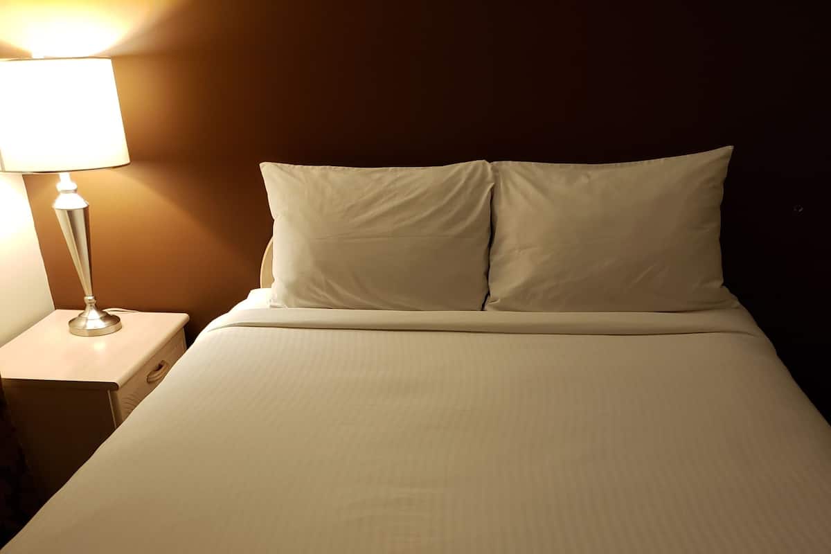 Una cama bien tendida con sábanas y almohadones blancos, una lámpara de noche y una mesita de madera.
