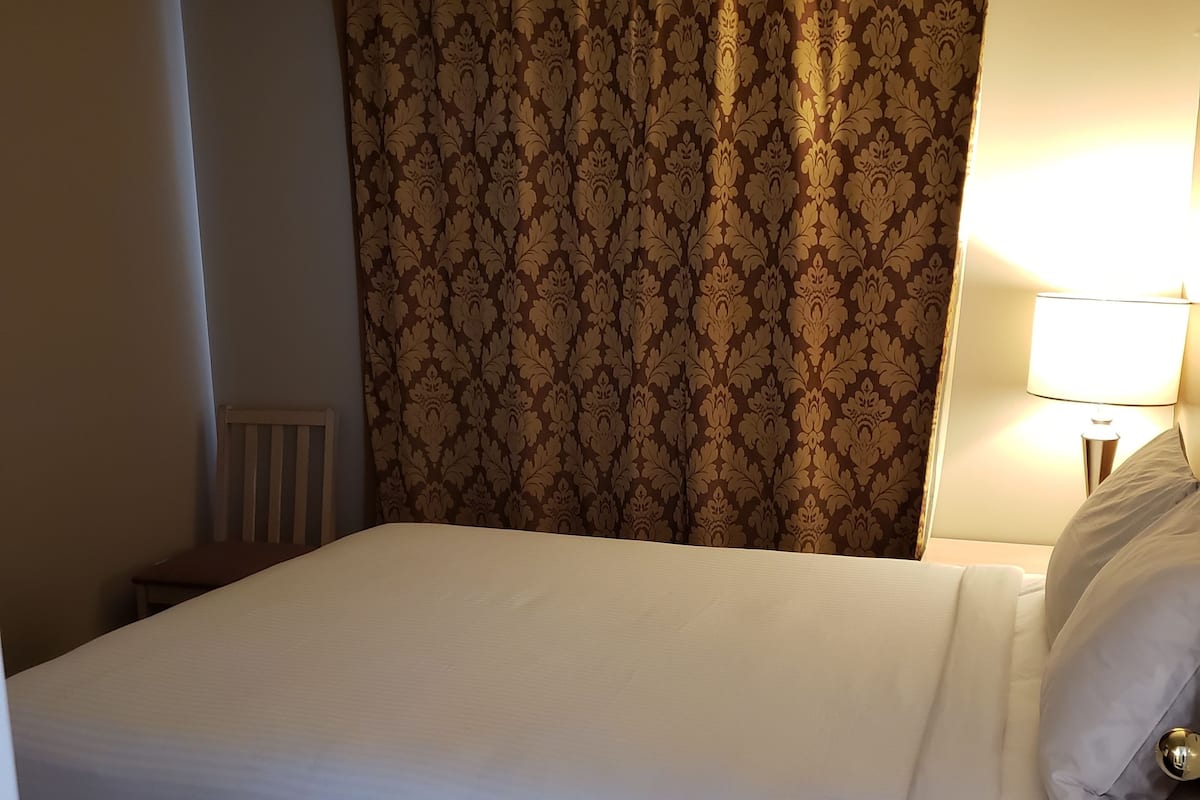 Una cama bien hecha con sábanas blancas, una cortina marrón estampada y una lámpara de noche.
